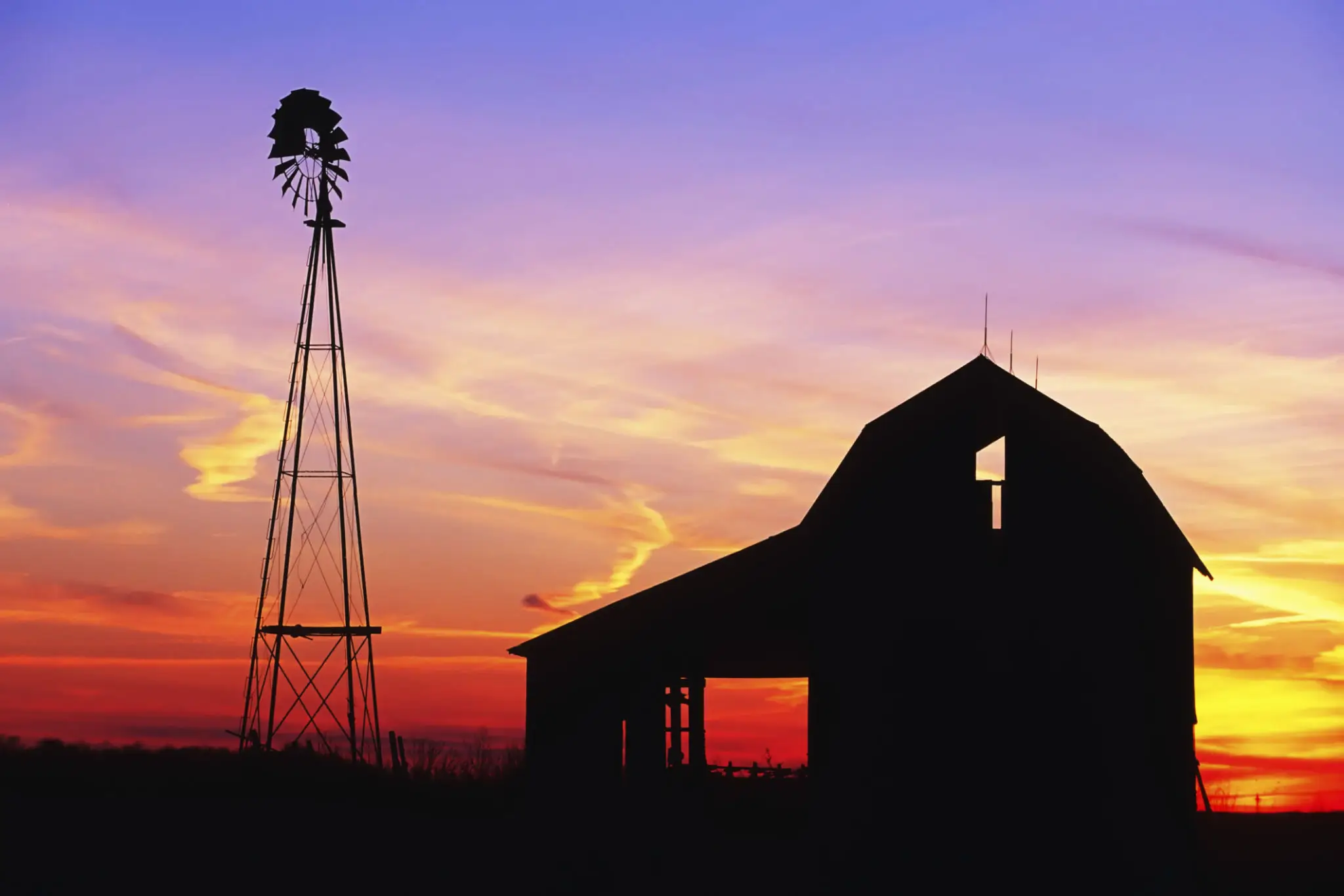 Silhouette of a barn and windmill against a vibrant sunset sky.