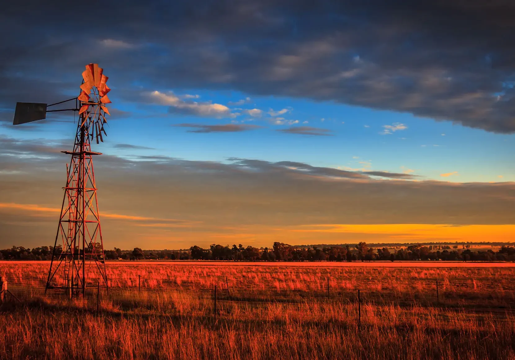 A windmill stands in a sunlit field under a colorful sky at sunset.